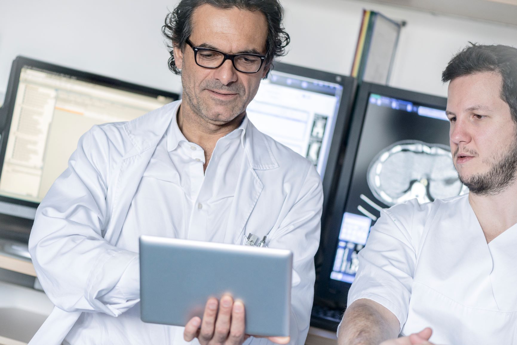 Bild von zwei männlichen Krankenhausmitarbeitern in weißer Arbeitskleidung vor Monitoren und mit einem Tablet in der Hand - Bildnachweis: Stock 